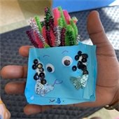 a child holding a folded piece of paper with a face drawn on it. inside the paper are pipe cleaners sticking out like colorful hairs.
